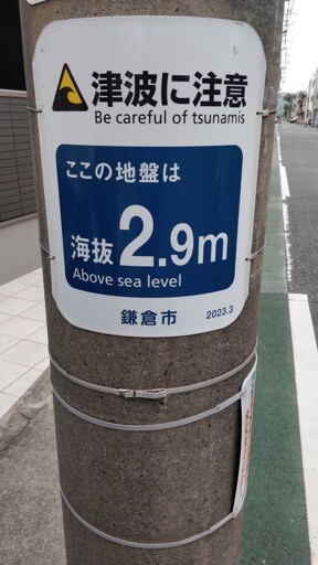 The photo shows a gray concrete pole with a sign in white and blue. The sign displays a tsunami warning in Japanese and English. The English translation reads: "Be careful of tsunamis." Below in the blue part of the sign there's an altitude information, also in Japanese and English, indicating the height above sea level. The English translation is "Above sea level," and the value is 2.9 m. At the bottom of the sign, the name of the city Kamakura and the date (2023.3) are shown in Japanese. The pole stands at the edge of a sidewalk. Other buildings, streets, and trees can be seen in the background.