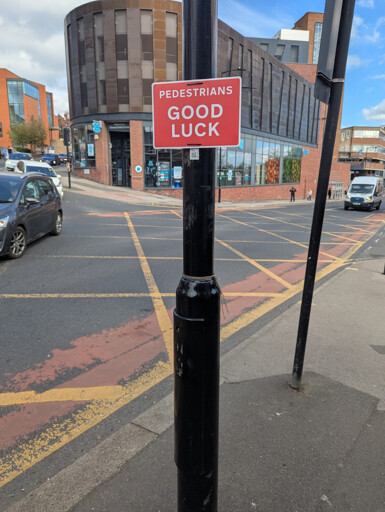 Picture of an official looking sign saying "PEDESTRIANS GOOD LUCK" attached to a lamppost in front of a busy intersection.