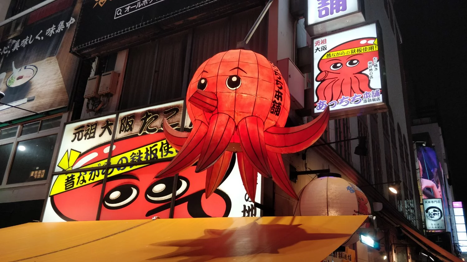 The photo shows a brightly lit street scene at night, in the Dotonbori area of Osaka, Japan. The most prominent feature in this picture is a large, three-dimensional Japanese lantern shaped like a red squid or octopus. The lantern is detailed with cartoonish eyes and a smiling expression. Its tentacles curve outwards and downwards. The lantern is illuminated from within, making it glow brightly against the darker backdrop of the surrounding buildings and night sky.
Behind and below the squid lantern, there are several brightly lit signs and billboards in Japanese. One prominent sign features a cartoon drawing of a red octopus with large eyes and the text "元祖大阪たこ焼" (Ganso Osaka Takoyaki), indicating it's advertising original Osaka-style takoyaki (octopus balls). Another sign above it also features a red octopus illustration and Japanese text.
The architecture suggests a bustling commercial area with multiple stories of buildings covered in various advertisements and signage, typical of entertainment districts in Japan. The overall atmosphere is vibrant and energetic, characteristic of Dotonbori's lively nightlife and street food scene.