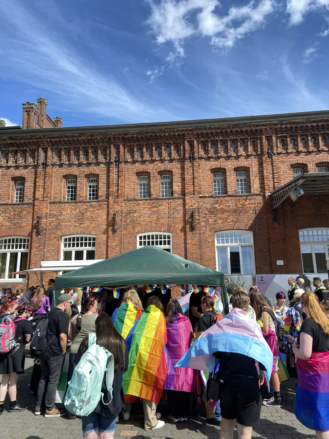 Besucher*innen des CSD mit LGBTIQ+ Flaggen auf dem Rücken am Stand für Sticker und Kram