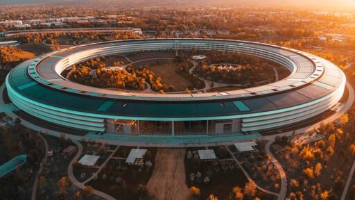 Aerial view of the Apple Park campus, showcasing its circular main building surrounded by landscaped grounds. The image captures the building's sleek, modern design with solar panels on the roof, set against a backdrop of trees and a sunset sky.