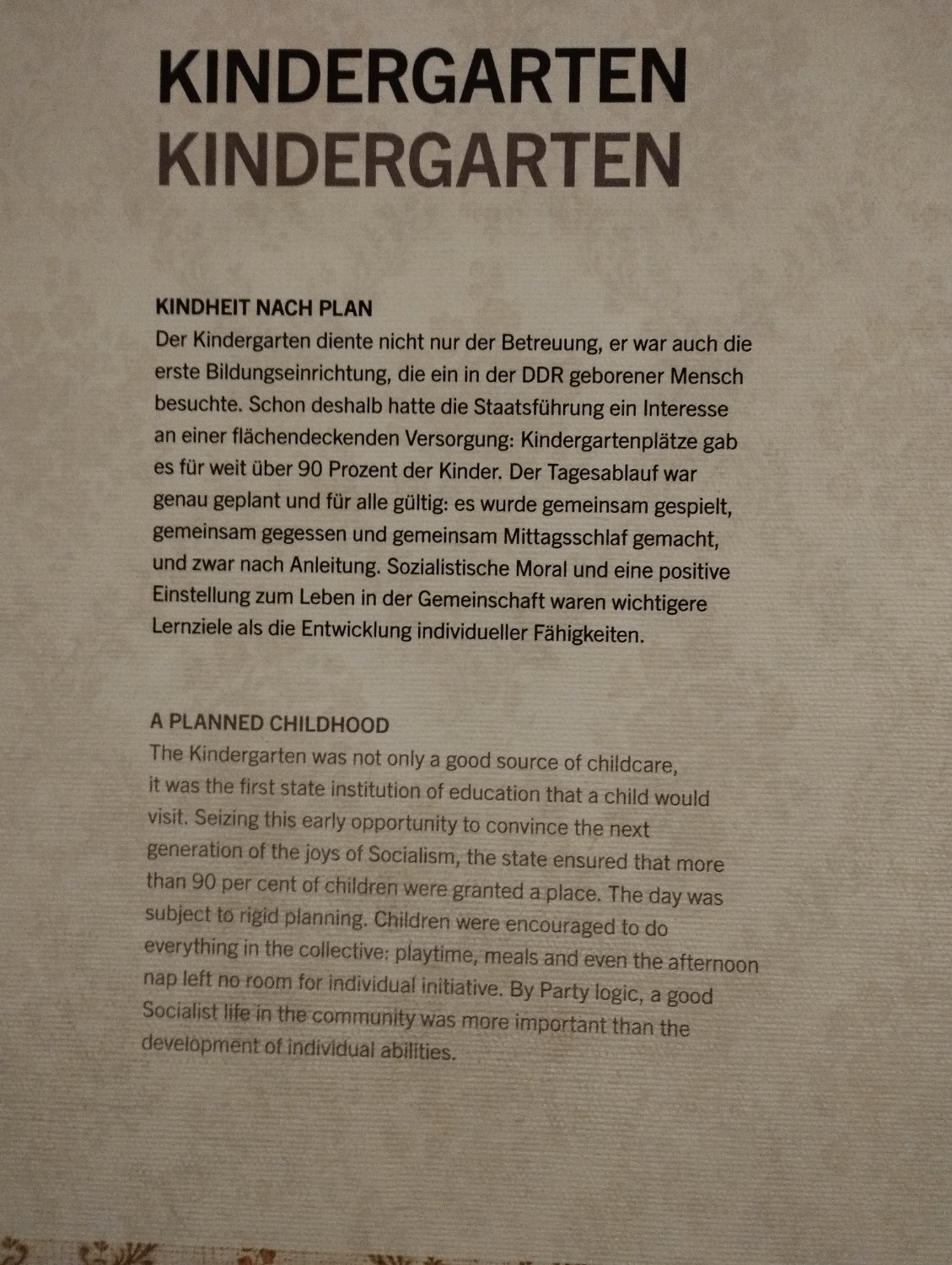 A note written in the wall of a museum saying: A PLANNED CHILDHOOD  The Kindergarten was not only a good source of childcare, It was the first state institution of education that a child would visit. Seizing this early opportunity to convince the next generation of the joys of Socialism, the state ensured that more than 90 per cent of children were granted a place. The day was subject to rigid planning. Children were encouraged to do everything in the collective: playtime, meals and even the afternoon nap left ne room for individual initiative. By Party logic, a good Socialist life in the community was more important than the development of individual abilities