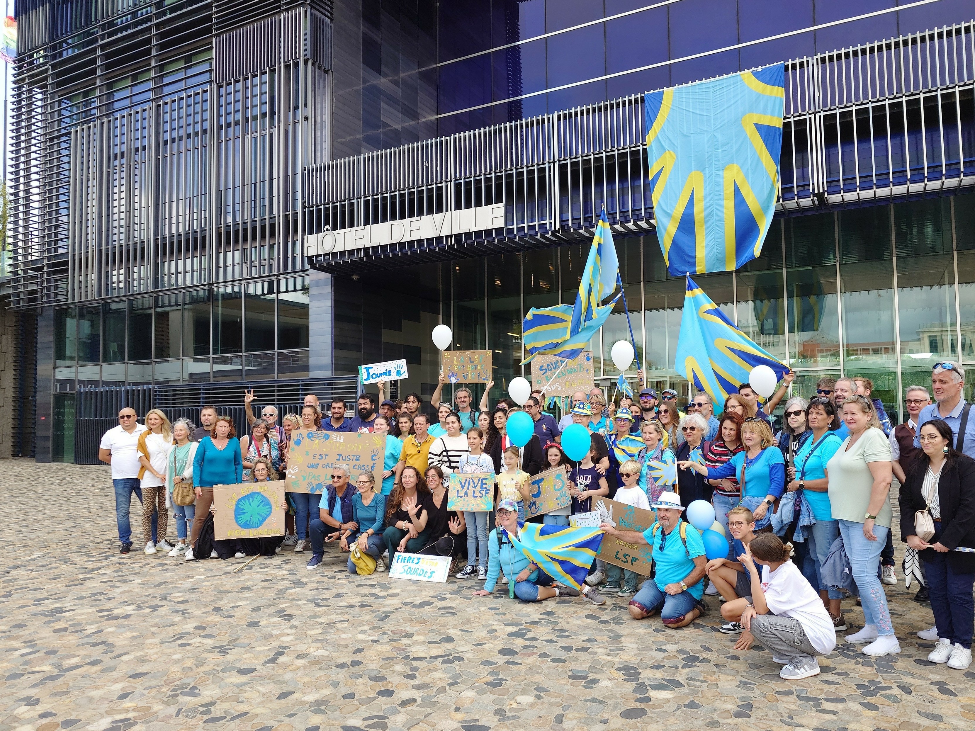 Photo de hamille avec les sourds et sourdes de montpellier groupés derrière le drapeau sourd accroché à l'hotel de ville