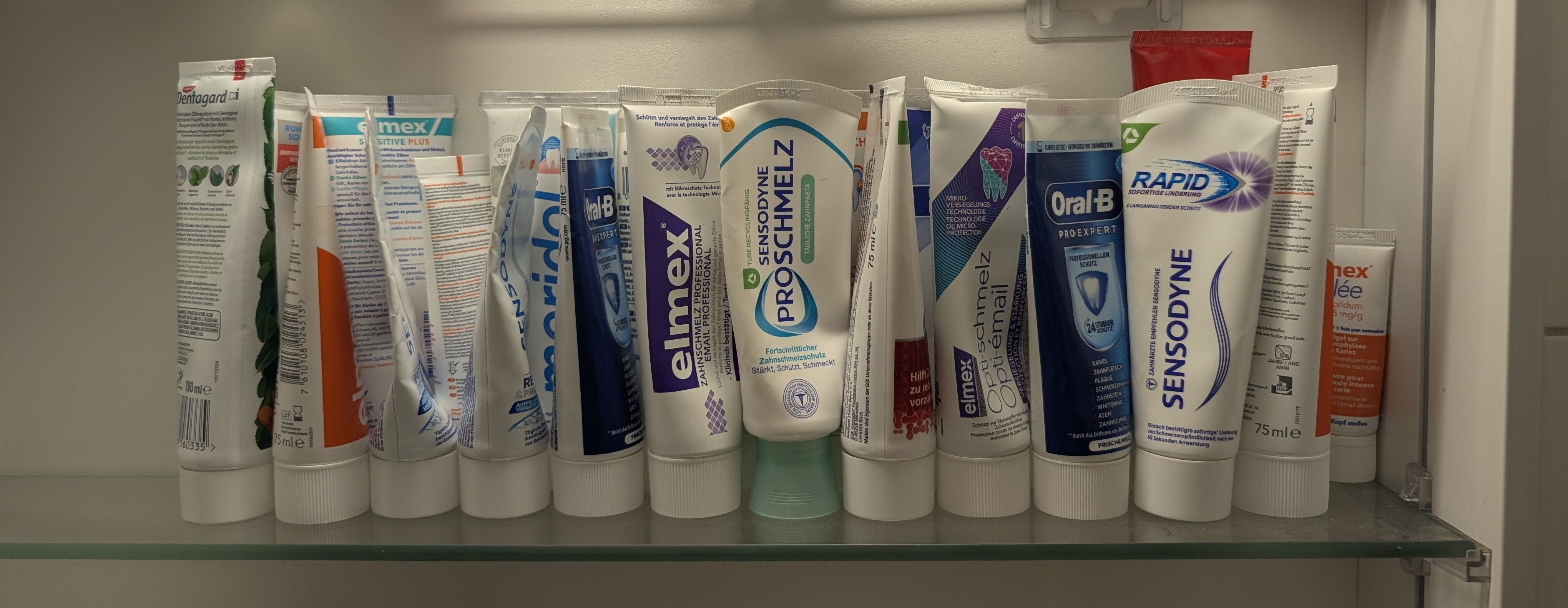 Two dozen toothpaste tubes arranged in two rows on a glass bathroom
shelf, featuring various brands with predominantly blue, white, and
green packaging, with no duplicated products. The tubes are upright
standing on their lids.