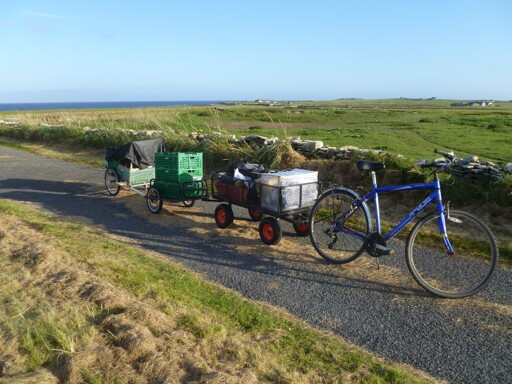 On a narrow road: A blue bicycle, with a train of 3 trailers connected behind it. Behind the bike are fields and the sea.