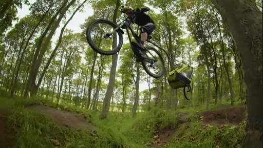 Mountain biker Danny McAskill performs a high jump on his bike in a forest setting, surrounded by trees, with a yellow trailer attached to his bike.