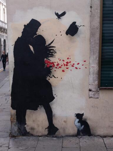 Questa è un murales di strada su un muro di un edificio. Il murale presenta silhouette nere di due figure: una vestita con un cappello, l'altra con lunghi capelli e un vestito, abbracciate in un bacio. Forme a forma di petali rossi si staccano dalla figura con i lunghi capelli. Un uccello nero con ali spiegate vola vicino in cima al murales, e un piccolo gatto nero e bianco si trova in fondo all'aspetto destro. L'edificio ha un muro con una parete texturizzata, di colore chiaro, con una tenda verde a destra e una persona cammina per una strada in pietra nel retro.