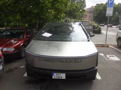 Tesla Cybertruck with custom near-invisibly rounded edges to pass EU standards, charging at a parking lot in central Prague. It is monstrous compared to a 2000s family car next to it. Giant "CYBERTRUCK.EU" sticker on the front. Licence plate: "001 CYBER". The weather is sunny but the car has been splashed with water, with a sheet of paper left on the windshield.