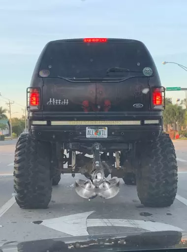 An image of a black sedan with a high lift kit, oversized tires, a Florida license plate reading “all evil”, and a pair of “truck nuts” that have been edited to be 2 demon cores.