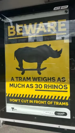 An PSA advertisement at a tram stop. In large text at the top is "BEWARE", then a pictograph of a rhino standing on a skateboard. Below reads "A tram weighs as much as 30 Rhinos" then below that again, reads "Don't cut in front of trams".