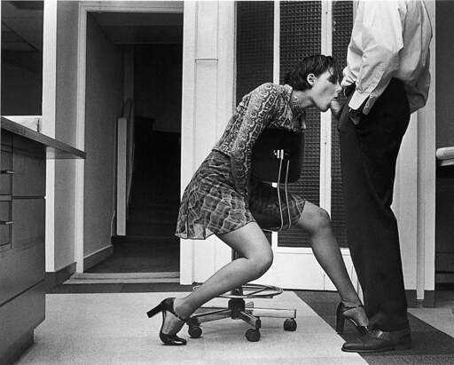 Black and white photo of Chloe Des Lysses gives a man a blowjob in an office setting. The mans head is out of frame. Both are wearing business attire