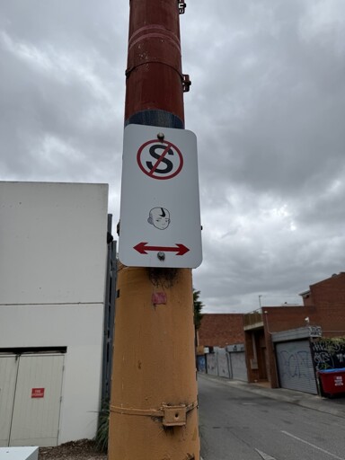A 'No Stopping' street sign attached to a red and yellow pole, with a sticker of a disappointed Aang from Avatar: The Last Airbender placed below the symbol. The arrow tattoo on Aang's forehead has been replaced with an rotated Nike swipe. The background shows an overcast sky, buildings, and a graffiti-covered alleyway.